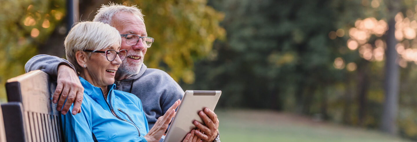 An older couple on a bench in a park looking happily at a tablet