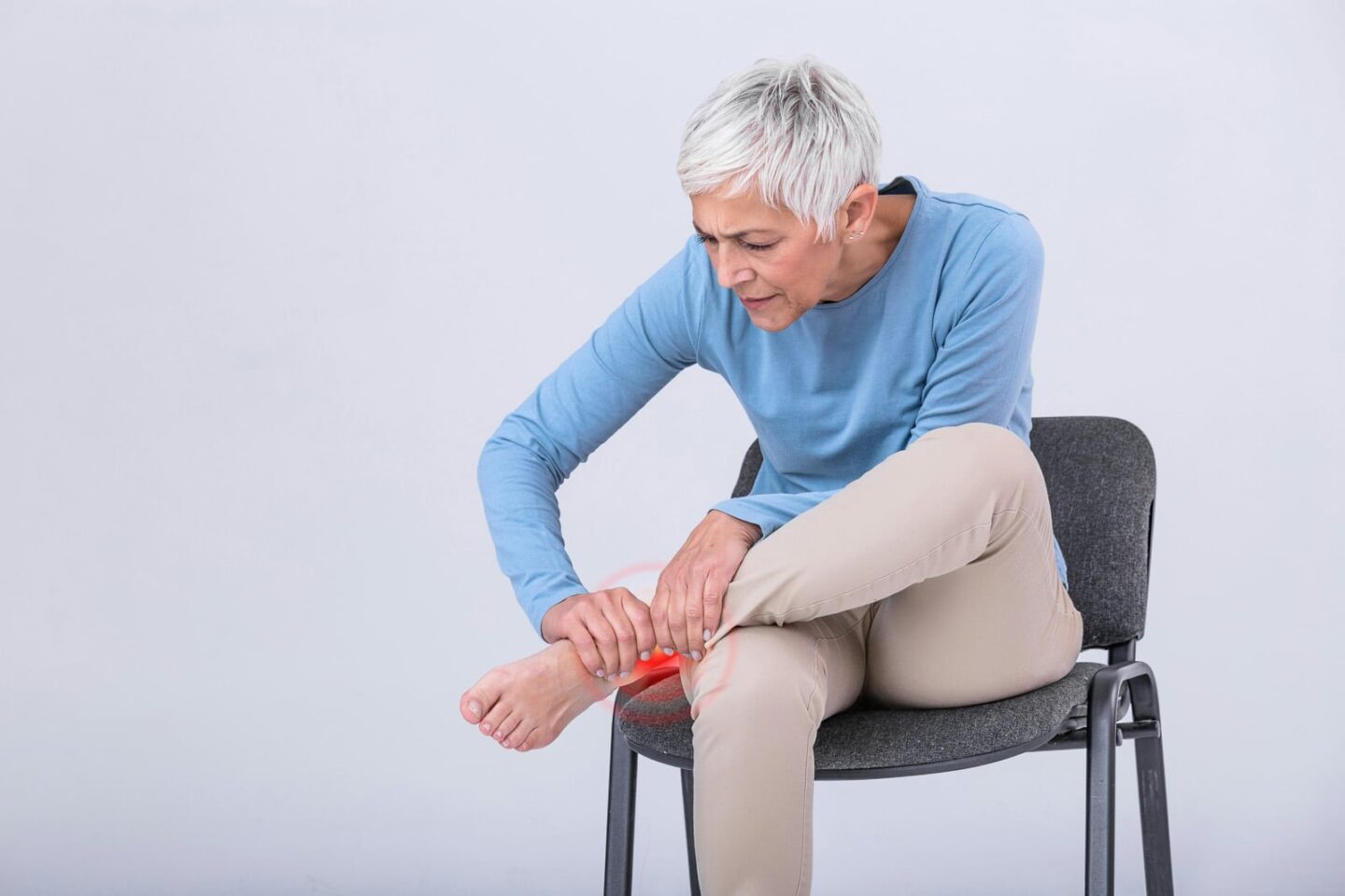 An older adult woman sitting in a chair wincing while she holds her ankle, red pain radiating