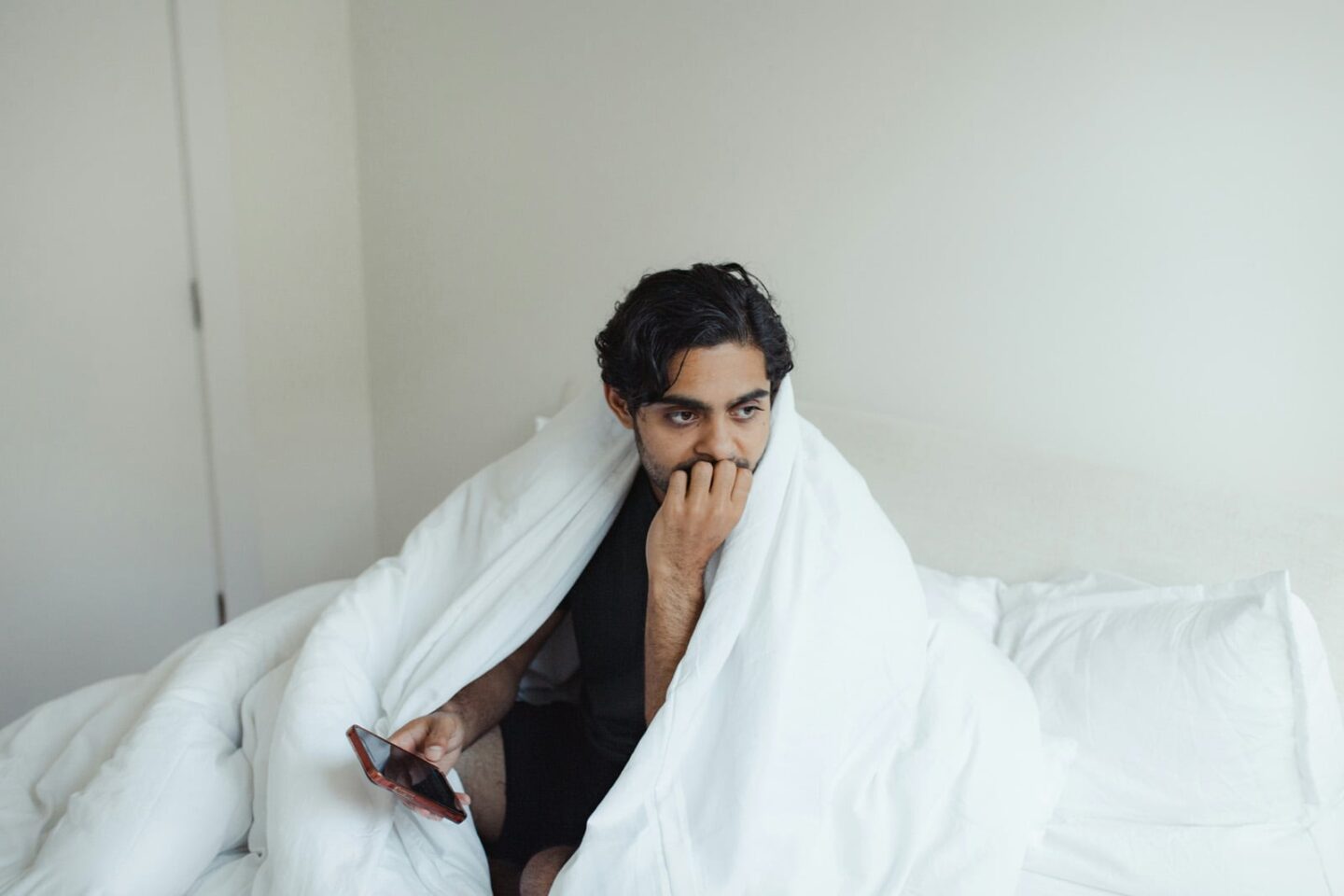 A man bundled in a blanket sitting on his bed, with a pondering look